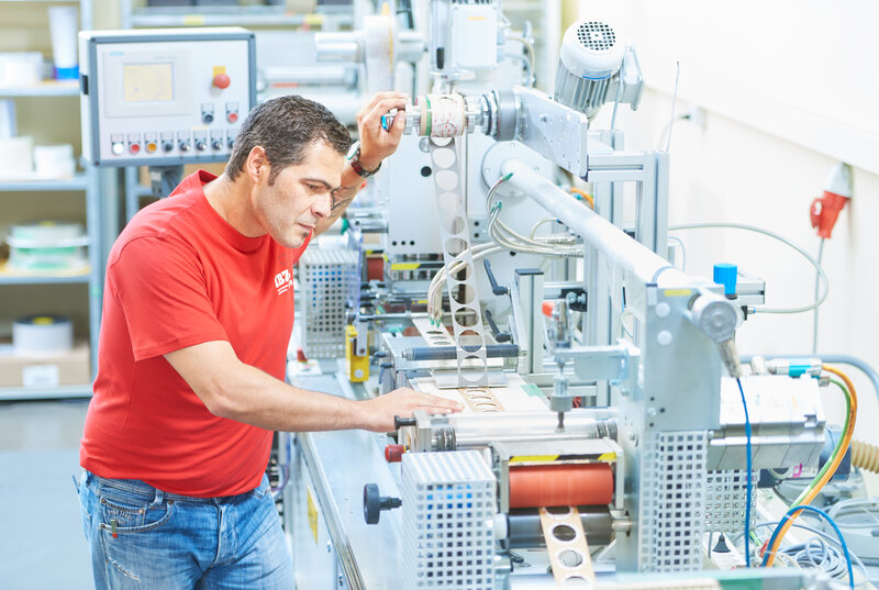 Auf dem Bild ist die Maschine für das Flachstanzen abgebildet, welche von einem Mann im roten IBZ-Shirt betrieben wird. Das gestanzte Band hat Kreise eingestanzt.