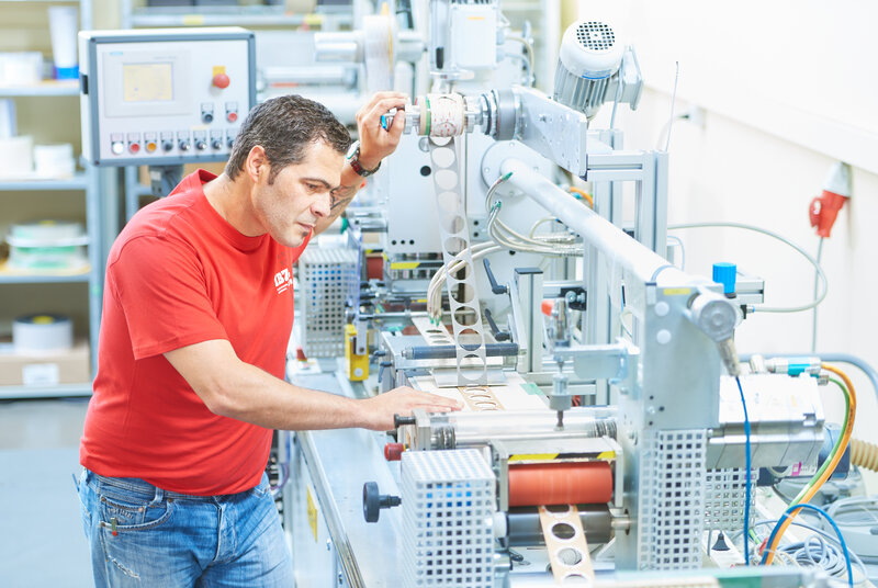 Abbildung Flachstanzen Auf dem Bild ist die Maschine für das Flachstanzen abgebildet, welche von einem Mann im roten IBZ-Shirt betrieben wird. Das gestanzte Band hat Kreise eingestanzt.