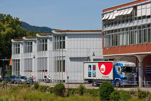 Camion IBZ à l'arrêt devant le bâtiment d'IBZ Industrie AG à Adliswil. Le camion est représenté devant la rampe de chargement. En bas de l'image, on peut voir la banlieue verte de la Sihl.