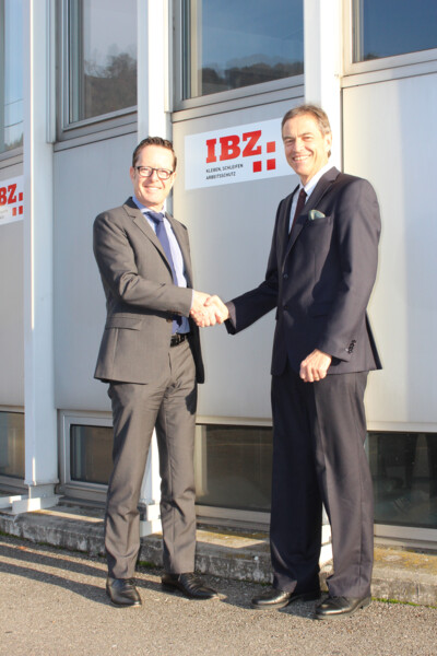 Remise des clés de l'IBZ Industrie AG entre Urs Egli et Roger Fehlmann. Les deux se tiennent devant le bâtiment de l'IBZ Industrie AG et se serrent la main.