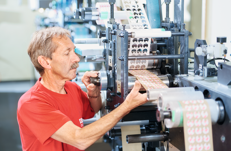 Image poinçonnage rotatif Sur l'image de la découpe rotative, on voit à gauche un employé en t-shirt rouge de l'ibz qui utilise la machine de découpe rotative. Sur la droite, on voit comment des cercles sont découpés dans la bande.