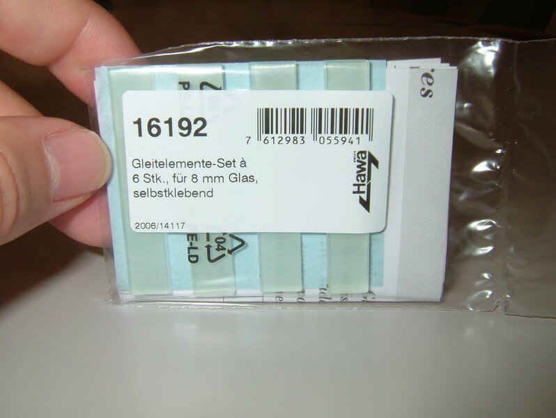 Die Abbildung zeigt eine Packung selbstklebende gleitelemente in sets à 6 Stück für 8 mm Glas. Die Verpackung ist durchsichtig  mit einem Barcode. die packung wird von einer hand gehalten und hat Blitzreflektionen