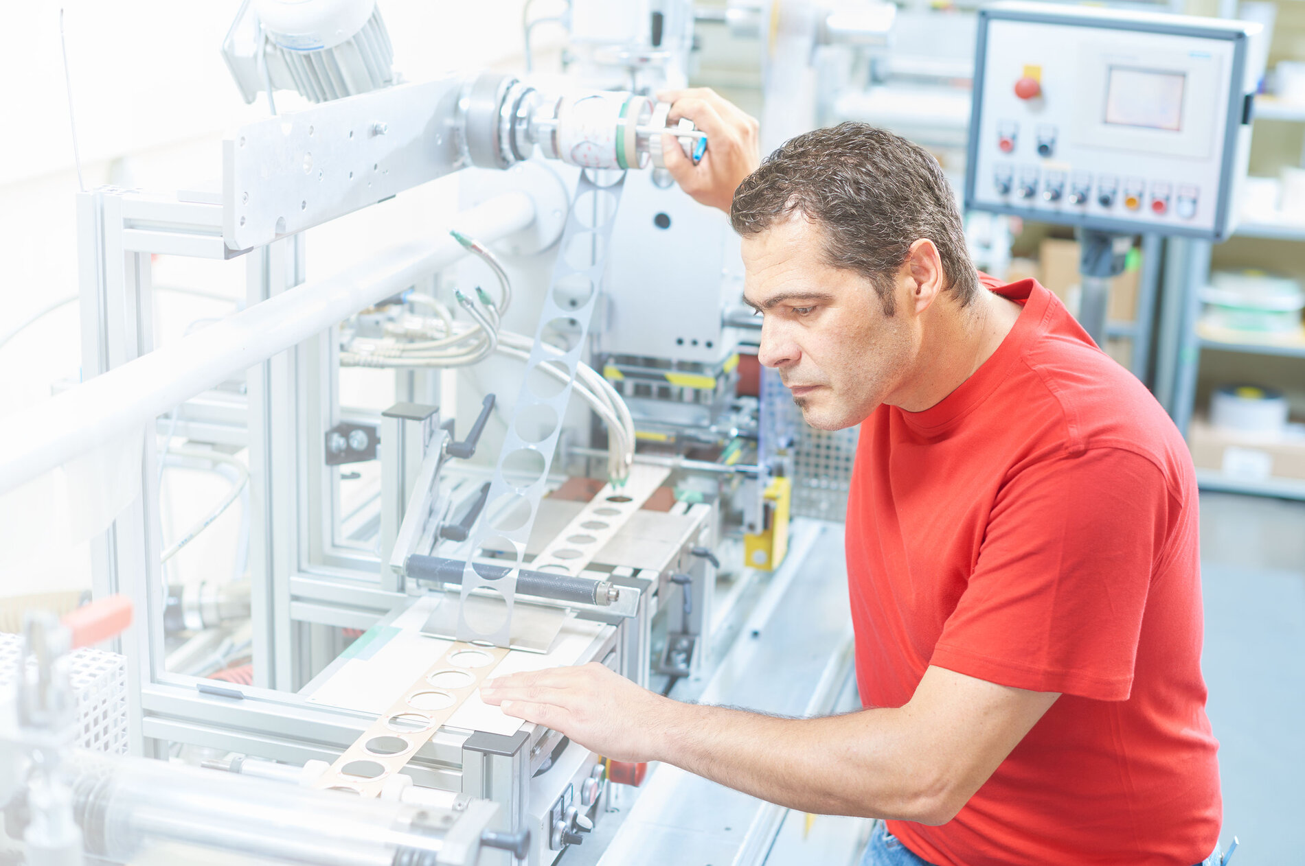 Auf dem Bild ist die Produktion von Formstanzteile durch eine Maschine, die von einem Arbeiter bedient wird, zu sehen. Dieser ist in einem roten IBZ Shirt. Die Maschine druckt runde Teile aus